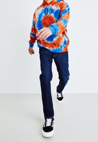 Sudadera tie-dye en naranja, azul y blanco; combinada con jeans azul oscuro y zapatillas negras. La sudadera cuenta con un bolsillo frontal y cordones.