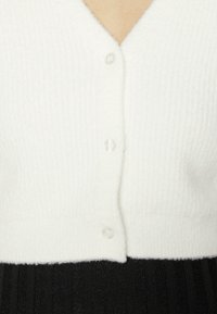 Bílý texturovaný cardigan s V-výstřihem a pěti průhlednými knoflíky. Měkký materiál, zkrácený střih s žebrovaným vzorem na lemu.