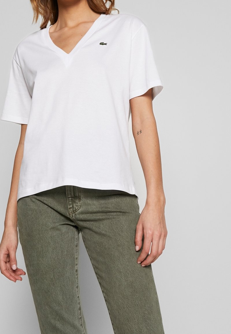 Kvinna iklädd vit T-shirt med V-hals och korta ärmar, prydd med en liten krokodil-logotyp, samt olivgröna jeans, stående mot en enkel grå bakgrund.