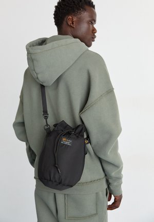 Sac à cordon noir en matériau durable, avec un écusson logo, porté en bandoulière avec un sweat à capuche vert clair.