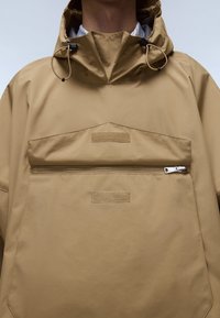 Jachetă bej impermeabilă, cu guler înalt, șnururi reglabile și buzunar frontal cu fermoar și clapetă. Textură netedă, design minimal.