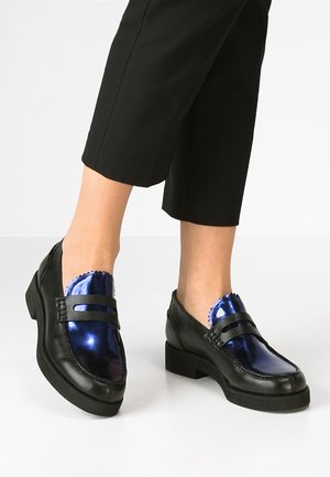 Füße in schwarzen Loafern mit glänzenden metallisch blauen Obermaterialien, kombiniert mit schwarzen, gekürzten Hosen vor einem schlichten Hintergrund.