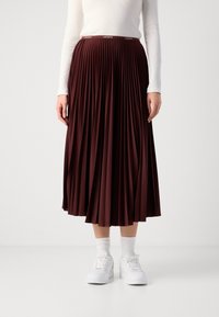 Jupe plissée bordeaux avec une ceinture élastique arborant le logo "Lacoste" à la taille, associée à des baskets blanches et des chaussettes de cheville.