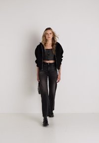 Črna zip-up kapuca, črtasta črna kratka majica in temne razširjene jeans hlače. Model drži majhno črno-belo torbico s pikami. Enostavna ozadje.