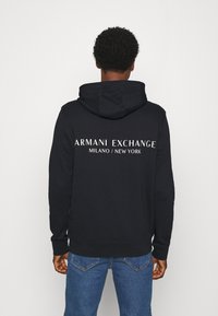Sudadera con capucha negra que presenta el texto "ARMANI EXCHANGE MILANO / NEW YORK" en la parte posterior. Confeccionada en tela suave, con un bolsillo tipo canguro y puños acanalados.