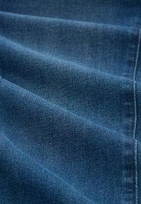 Tejido de mezclilla en tono azul profundo, mostrando una textura suave con un ligero desgaste, cayendo en pliegues y con costuras visibles a lo largo de los bordes.