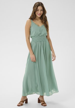 Femme en robe maxi sans manches vert clair avec des détails en dentelle, souriante, debout, portant des sandales à talons marron sur un fond blanc.