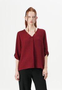 Blusa bordeaux con scollatura a V, tessuto testurizzato, maniche a tre quarti con risvolto e vestibilità rilassata. Abbinata a pantaloni neri.
