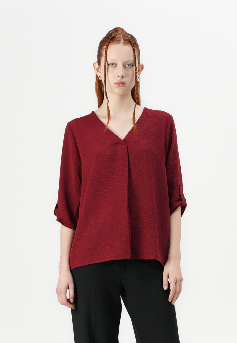 Blusa bordeaux con scollatura a V, tessuto testurizzato, maniche a tre quarti con risvolto e vestibilità rilassata. Abbinata a pantaloni neri.