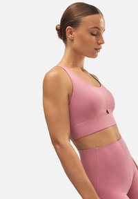 Sostenedor deportivo rosa hecho de tejido suave, con escote redondeado, tirantes anchos y una banda debajo del busto con una pequeña abertura en el centro.