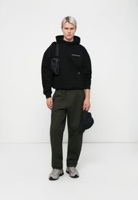 Zwarte hoodie met tekst, olijfgroene cargo broek, grijze sneakers en zwarte pet. Draagt een kleine zwarte schoudertas.