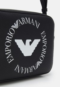 Black leather bag featuring white Emporio Armani logo and eagle emblem, rectangular shape, textured surface, and adjustable strap.