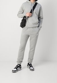 Osoba v šedé mikině Nike a teplákách, s černou krosovou taškou a černo-bílými teniskami Nike stojí na jednobarevném pozadí.