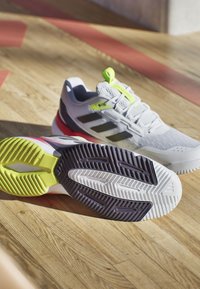 Białe buty sportowe z szarymi akcentami, czarnymi paskami i jaskrawozielonym piętą. Chropowata podeszwa w zielonym, purpurowym i białym kolorze.