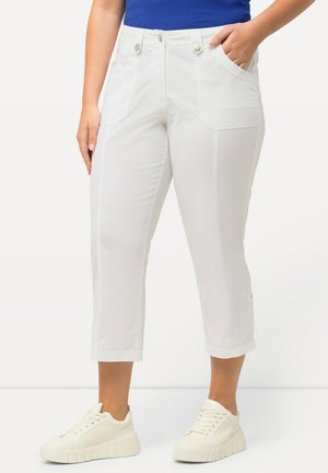 Femme portant un pantalon blanc court avec poches avant et des baskets blanches à plateforme, habillée d'un haut bleu, debout avec la main dans la poche.