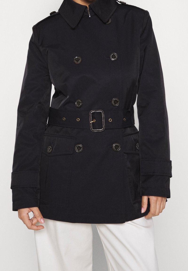 Dubbelknäppt trench coat i marinblått. Tillverkad av textil med struktur, har bälte i midjan, stora knappar och framfickor.