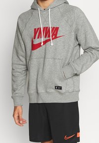 Personne portant un sweat à capuche gris avec le texte rouge "YNWA" et un logo swoosh, un short noir avec un swoosh Nike orange, main dans la poche du sweat.