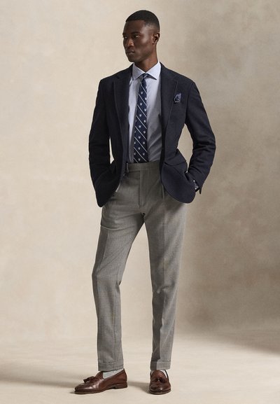 Mann trägt marineblauen Blazer, hellblaues Hemd, gemusterte Krawatte, graue Hose und braune Tassel-Loafer, steht mit Händen in den Taschen.