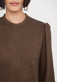 Haut marron à motif chevron texturé avec un col rond et des manches bouffantes, mettant en valeur un motif de tissage subtil avec une finition lisse.