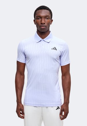 Homme portant un polo à manches courtes violet clair et un pantalon blanc, debout droit devant un fond clair uni.