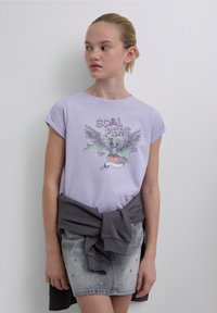 Lila T-shirt met korte mouwen, voorzien van een afbeelding van een grijze adelaar en tekst. Gecombineerd met een grijze denim rok versierd met strass-steentjes.