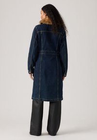 Manteau en denim lavage foncé avec col en fausse fourrure, taille ceinturée et manches longues. Présente des coutures contrastantes et des poches latérales.