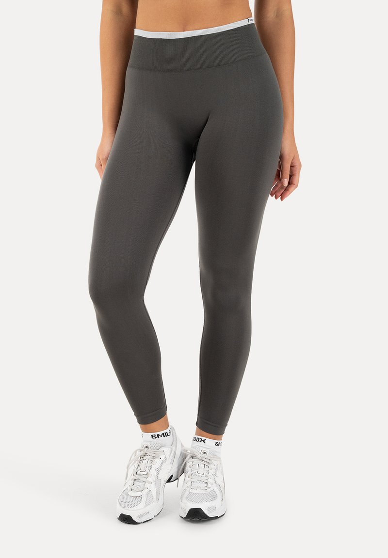 Femme portant des leggings gris foncé taille haute et des chaussures de sport blanches avec des chaussettes blanches assorties, debout devant un fond uni.