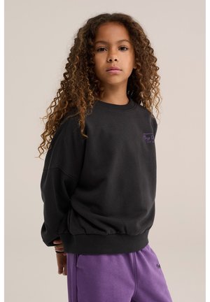 Jeune fille aux longs cheveux bouclés portant un sweat-shirt noir et un pantalon violet, debout devant un fond beige uni.
