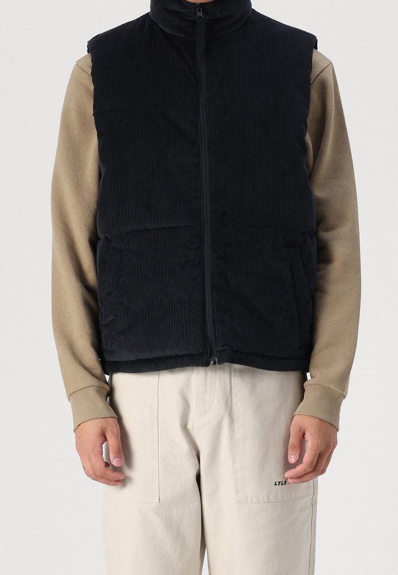 Chaleco de pana negro con cuello alto, cremallera frontal y bolsillos laterales. Lleva puesto encima de una camisa de manga larga beige y pantalones color crema.