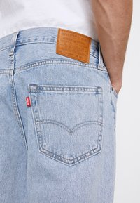 Jasnoniebieskie dżinsy z denimu z brązową skórzaną naszywką z napisem "Levi Strauss & Co." oraz czerwoną metką na tylniej kieszeni. Klasyczny design z pięcioma kieszeniami.