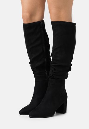 Bottes en daim noir montant jusqu'aux genoux avec des tiges souples, des fermetures éclair latérales, des bouts pointus et des talons blocs, portées sur des jambes nues sur un fond blanc.