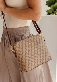 Beige vzorovaná kabelka s hladkým povrchem, zlatými prvky a hnědým popruhem. Taška má obdélníkový tvar a jemný detail loga.