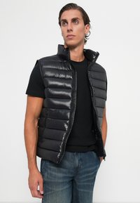 Bărbat cu păr închis la culoare, purtând o vestă fără mâneci, căptușită, de culoare neagră, peste un tricou negru și blugi albaștri, stând în fața unui fundal alb simplu.