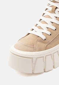 Shoe sneaker de camurça bege com sola de borracha branca robusta, apresentando costuras proeminentes e um design de atacadores com atacadores brancos.