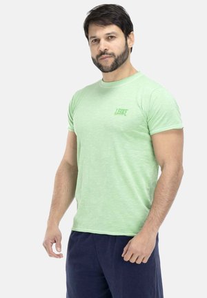 Mann mit Bart, der ein hellgrünes kurzärmeliges T-Shirt und dunkelblaue Hose trägt, steht vor einem einfarbigen hellen Hintergrund.