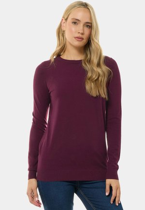Femme aux longs cheveux blonds portant un pull moulant à manches longues violet foncé et un jean bleu, debout devant un fond uni.