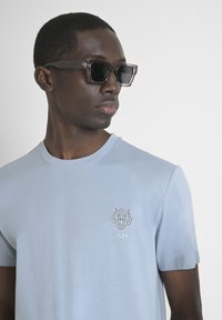 T-shirt in cotone azzurro chiaro con colletto rotondo, caratterizzato da un logo di tigre ricamato bianco sul lato sinistro del petto. Gli occhiali da sole hanno una montatura scura.