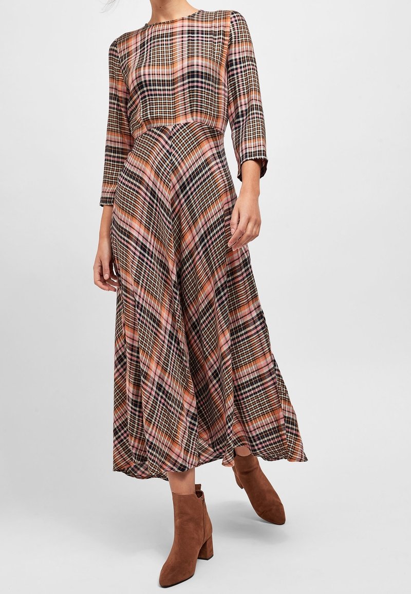 Femme portant une robe midi à manches longues à carreaux multicolores, associée à des bottines en suede marron sur un fond uni.