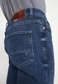 Tmavě modré džíny z denimu s hnědou koženou zadní nášivkou s ražebným logem. Na zadním kapse je malý detail ve formě barevné vlajky.