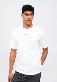 Hombre con cabello rizado que lleva una camiseta blanca con el texto negro "SAMSØE SAMSØE" y jeans oscuros grises, de pie contra un fondo claro y liso.