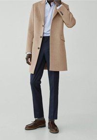 Beige wollen overcoat met een enkel-breasted ontwerp, gecombineerd met marineblauwe broek en bruine schoenen. Beschikt over ribbelmanchetten en kraag.