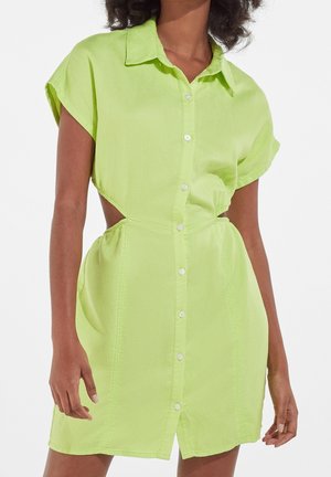 Mujer con un vestido camisa de manga corta color lima con los costados abiertos y cuello, de pie sobre un fondo blanco.