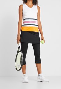 Ung kvinna som håller en tennisklubba och boll, klädd i en ärmlös vit topp med ränder, svart kjol med leggings och vita sneakers.