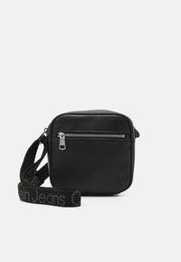 Črna usnjena torba čez ramo z zadrgo na sprednjem žepu, nastavljiv trak z napisom "Calvin Klein Jeans", kvadratne oblike, gladka tekstura.