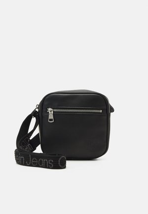 Borsa a tracolla nera in pelle con tasca anteriore con zip, tracolla regolabile con testo "Calvin Klein Jeans", forma quadrata, texture liscia.