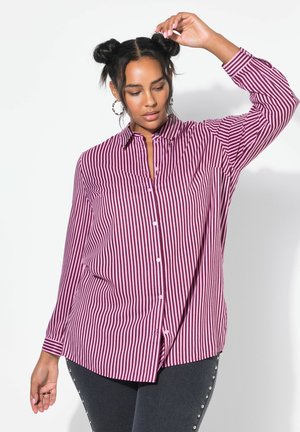 Camicia a righe bordeaux e bianca, con colletto, maniche lunghe e orlo spaccato, abbinata a jeans scuri con borchie.