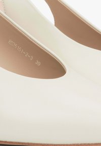 Bílé kožené lodičky na vysokém podpatku s elegantním designem, otevřenými boky a hnědou podrážkou. Uvnitř je vyražená velikost a kód stylu v jemném tisku.