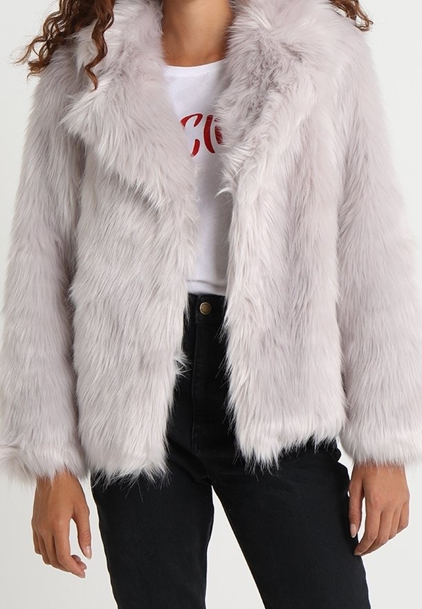 Femme portant une veste en fausse fourrure gris clair sur un t-shirt blanc à motif et un pantalon noir taille haute.