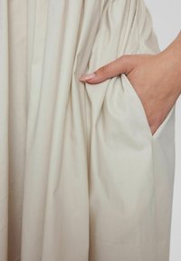Helles beiges Stoffkleid mit gefältelter Textur; verfügt über eine Seitentasche, aus der eine teilweise sichtbare Hand hervorkommt, die glatte Haut und gepflegte Nägel zeigt.