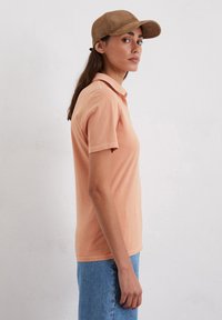 Perzikkleurige polo met korte mouwen, met een klassieke kraag en zijsplitten, gecombineerd met denim shorts en een bruine pet.